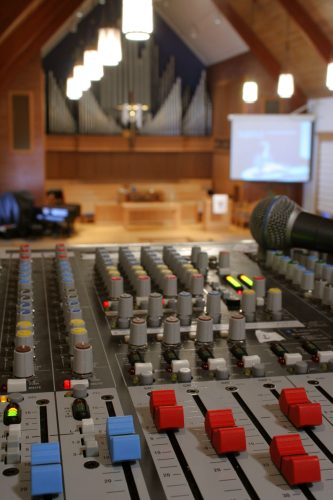 Peaceful view of a church sanctuary in soft focus behind a view of a sound/audio mixing board (console) and microphone.  Projection screen in background, with organ pipes and cross showing that it is a church. very sharp focus on the nearest sound board controls, with the service blurred by depth of field, though the center of the composition is the service & cross, not the tech.